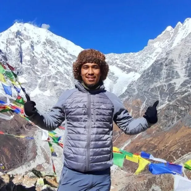 A trekker in a down jacket giving two thumbs up among prayer flags below snow-covered peaks